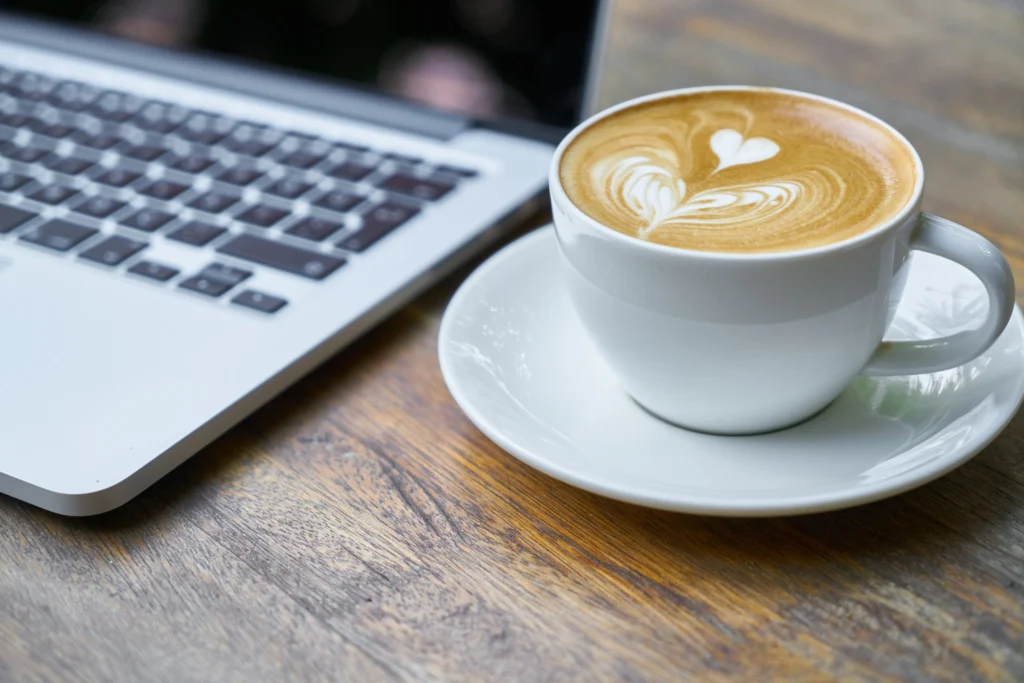 Sydney coffee guide showcases perfect latte with heart-shaped foam art on saucer next to laptop at Sydney cafe workspace similar to Cafe C&M, a favourite among Peakhurst Coffee Shops