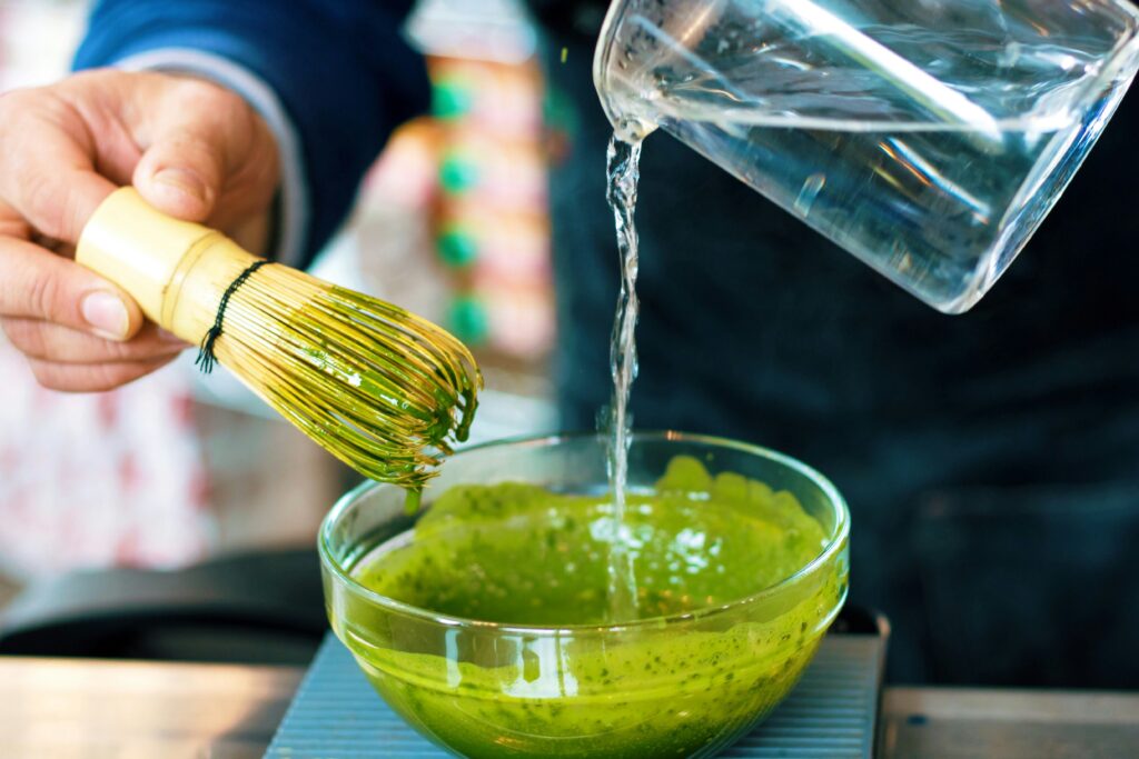 Barista preparing matcha using a traditional bamboo whisk while pouring hot water into a glass bowl filled with vibrant green matcha powder for an iced matcha drink at Cafe C&M Sydney.